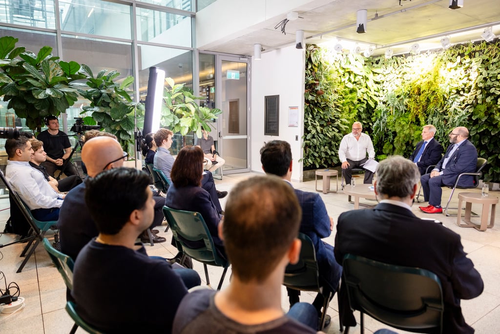 A group of students sit in chairs facing a small panel discussion set in a bright indoor space with large plants and a vertical garden wall. Three speakers sit at the front under studio lighting while attendees listen from rows of seats.