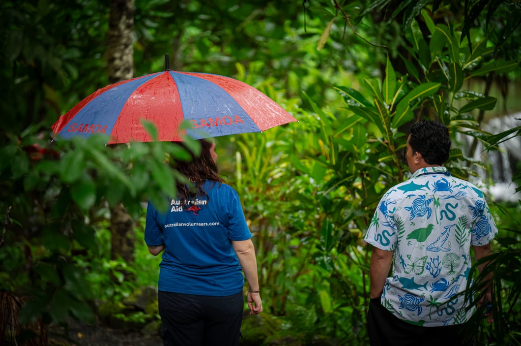 A botanist and a Senior Ranger from the Ministry of Natural Resources and Environment stand in the rainforest of Apia in Samoa. The woman on the left is holding a red and blue umbrella with a blue "Australian Volunteers" branded shirt. The man on the right is in a white shirt with a blue and green animal pattern. They are looking away from the viewer towards the plants in front of them.