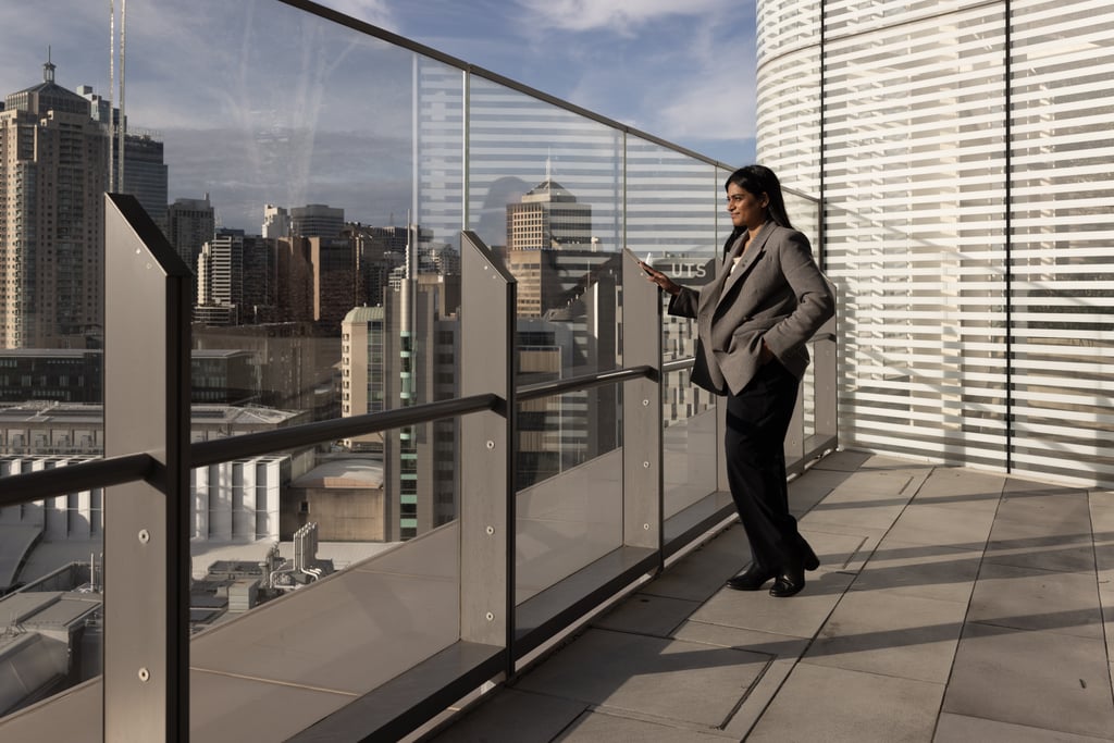 Ash stands on a balcony looking at her phone, with the Sydney skyline visible in the background