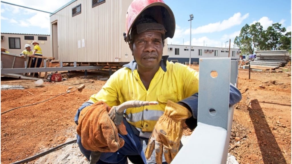 A construction worker in a yellow safety uniform and helmet crouches on a construction site, holding a tool. Mobile buildings and blue sky in the background.