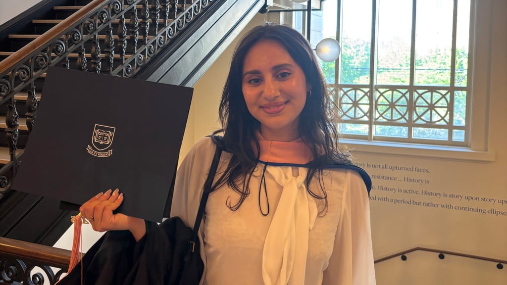 Jaya at her graduation, standing on an indoor staircase. She wears a white dress.