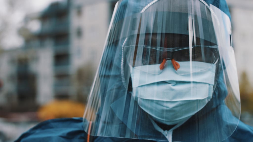 Person in protective gear, including a blue hood, face mask, and clear shield, stands outdoors. Background shows a blurred building, conveying a serious tone.