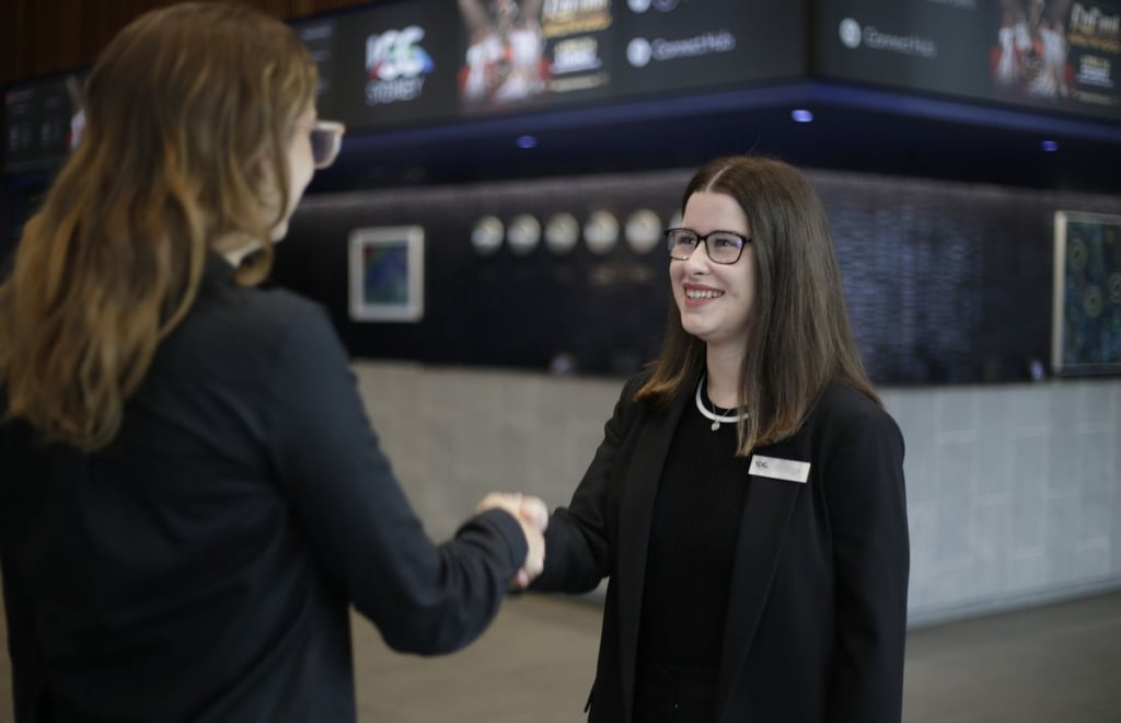 Emily Howe, a woman dressed in a black blazer and shirt, smiles as she shakes hands with an event attendee who has their back to the viewer. She wears glasses and has the ICC name tag attached to her blazer, in the background is the interior of the convention centre.