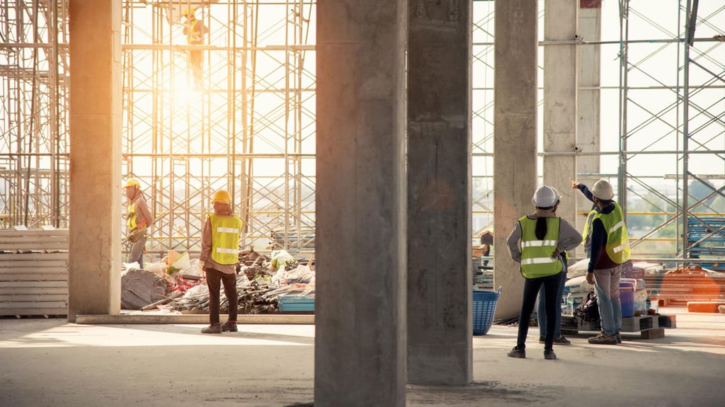 Construction site with workers in safety vests and helmets. They're standing near scaffolding, with sunlight streaming through, creating a focused and industrious atmosphere.