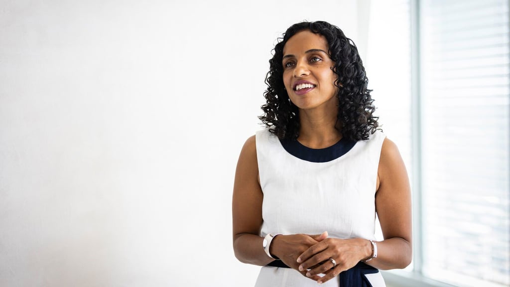Professor Ramona Vijeyarasa wears a white dress with black trim and stands in front of a window with blinds