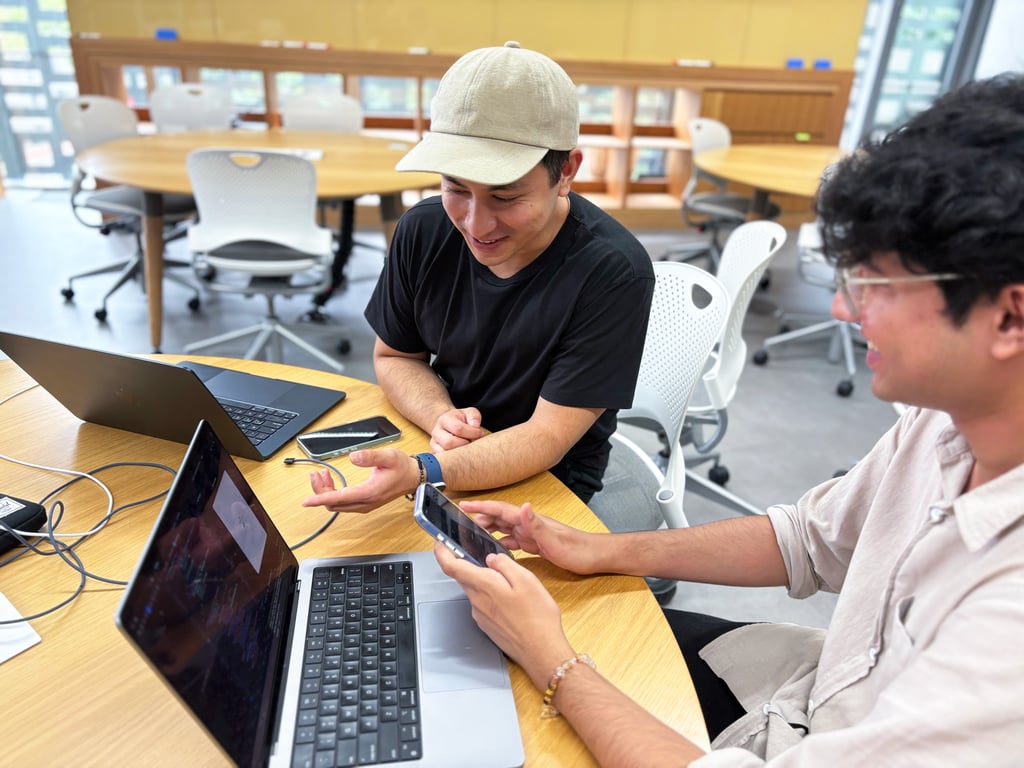 Two young men sit at a round table with laptops, smiling as they discuss something on a smartphone. The room has a collaborative, modern feel.