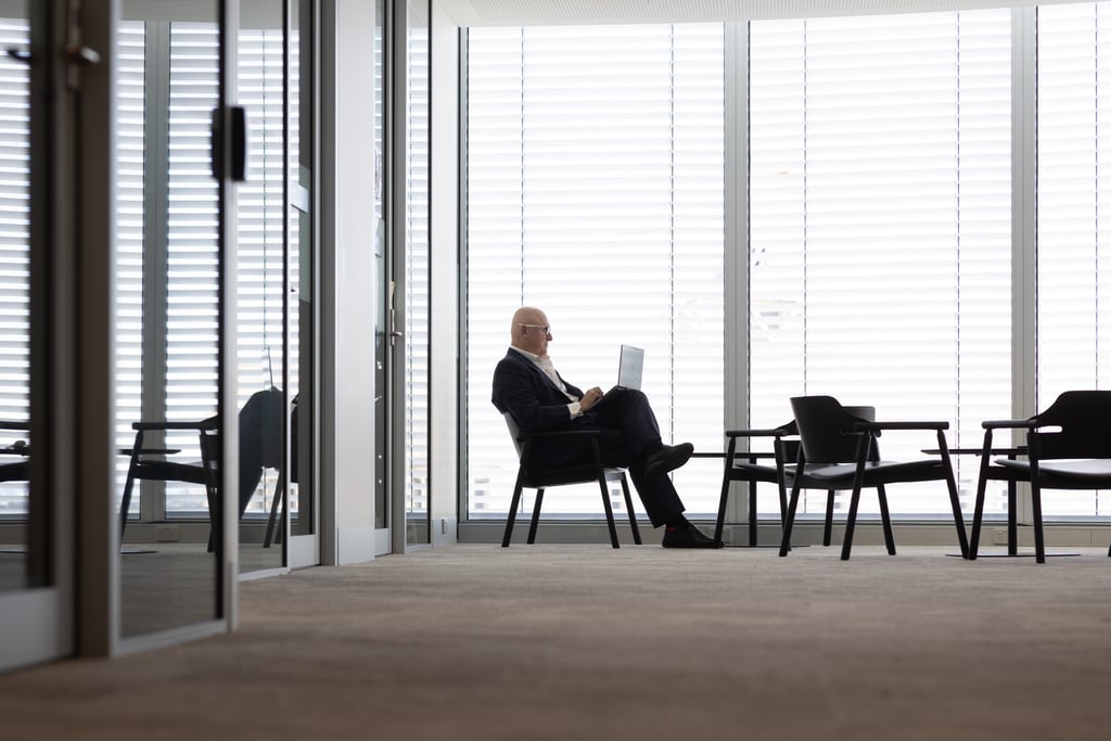 Nicholas sits in the distance, working on his laptop, in front of a window with while blinds