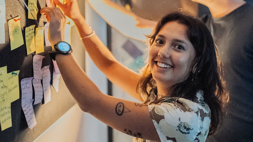 Two people place sticky notes on a wall covered with notes and connected materials during a collaborative planning session.