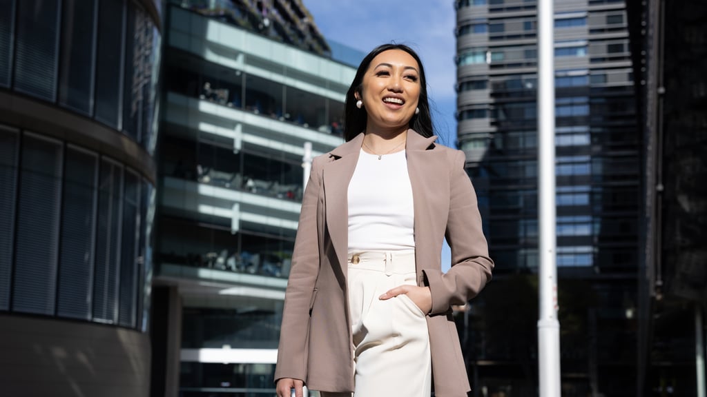 Arhita in a light brown blazer, smiles, standing in front of office buildings
