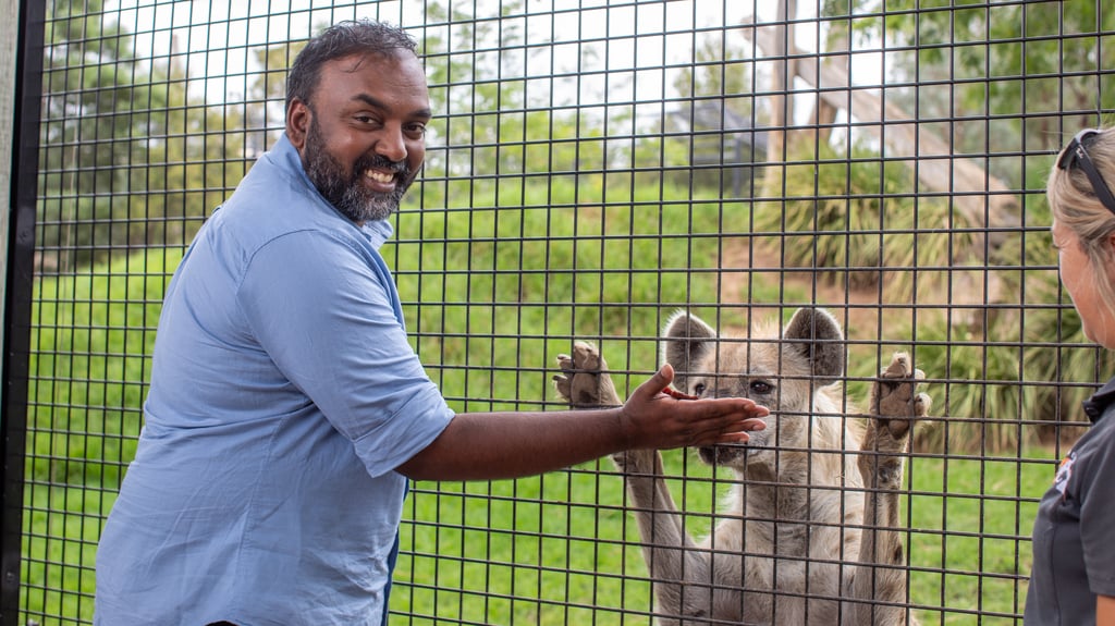 Hubert is dressed in a blue shirt and smiling to the camera. He reaches a hand up to a wired fence toward a spotted hyena standing upright with its front paws and nose against the mesh. Another person stands nearby observing, with green grass and zoo-style enclosures in the background.