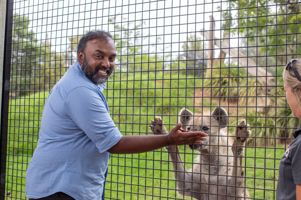 Hubert is dressed in a blue shirt and smiling to the camera. He reaches a hand up to a wired fence toward a spotted hyena standing upright with its front paws and nose against the mesh. Another person stands nearby observing, with green grass and zoo-style enclosures in the background.