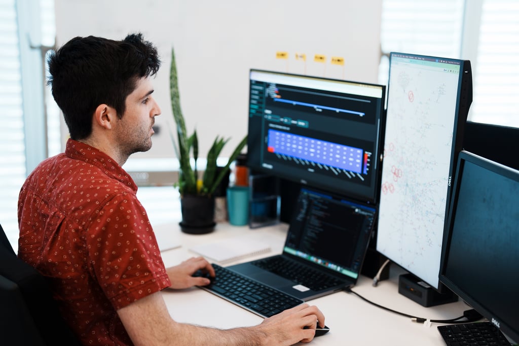 Software engineer reviewing data visualisation dashboards and a network diagram across multiple monitors and a laptop.