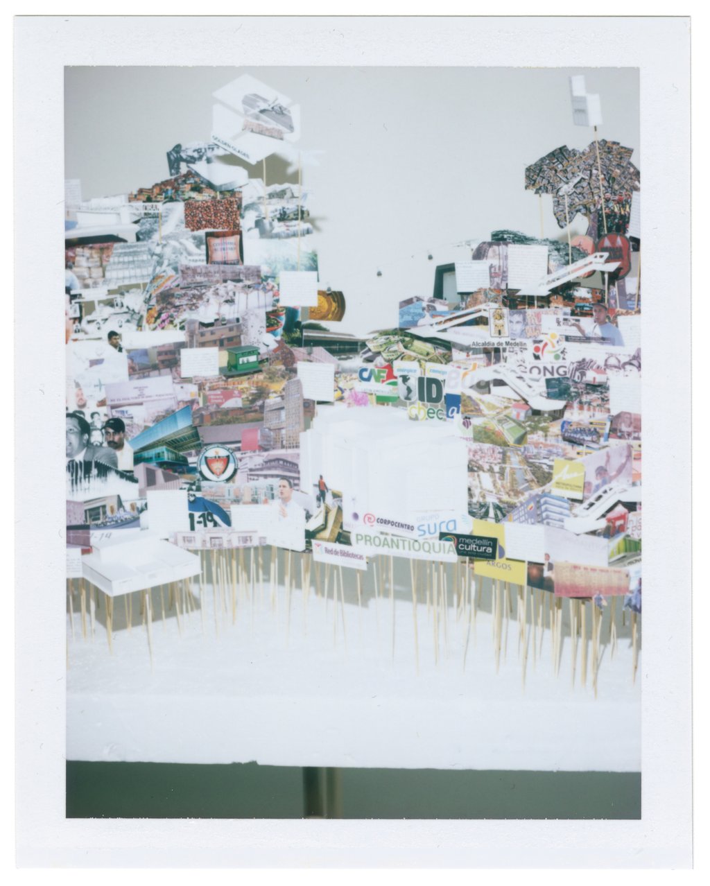 A framed photograph of part of the art installation representing the city of Medellin consisting of pictures on paper and folded paper artefacts mounted on sticks of varying heights and embedded in a flat white base.