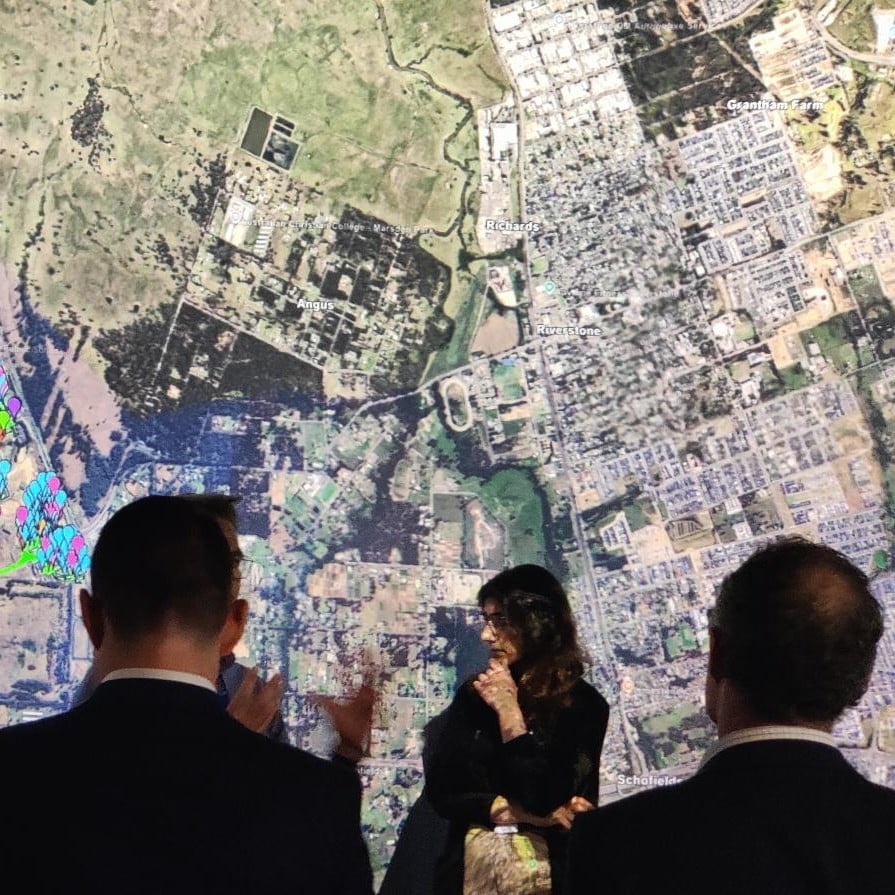 Group of people observing a large, detailed aerial map with highlighted zones in blue, green, and pink. The scene conveys engagement and collaboration.