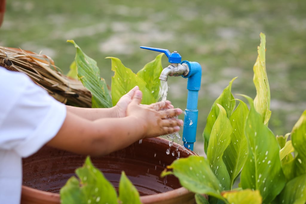 A child washes hands under a blue faucet, surrounded by lush green leaves.