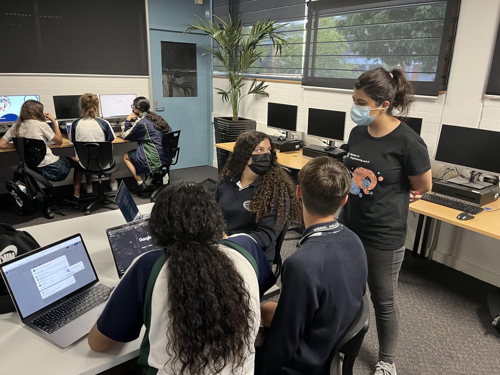 A classroom setting with students working on laptops. One student stands, wearing a mask, engaging with three others seated at a table. The mood is focused.