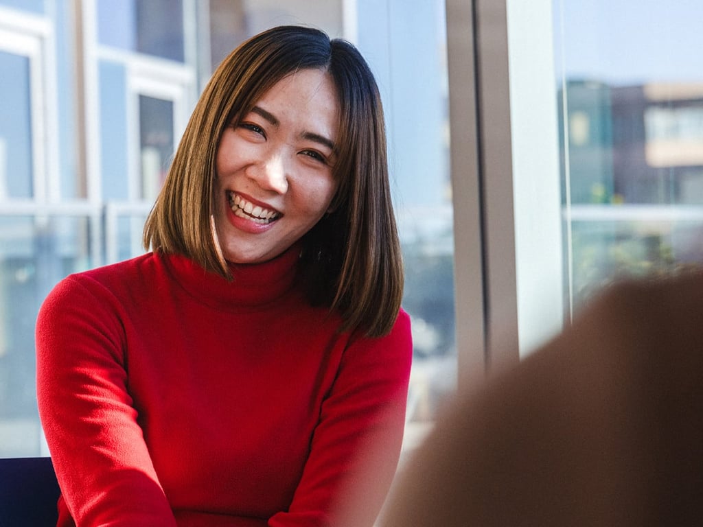 A woman in a red sweater smiles warmly while sitting indoors. A man nearby looks at his phone, laughing. Sunlight streams through large windows, creating a cheerful atmosphere.