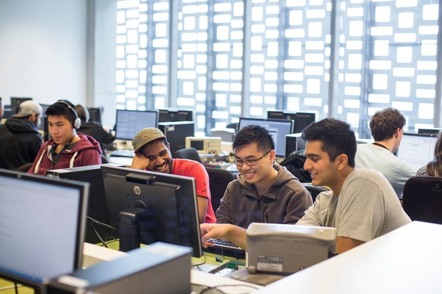 Students work together in a computer lab, seated at desktop computers arranged in rows.