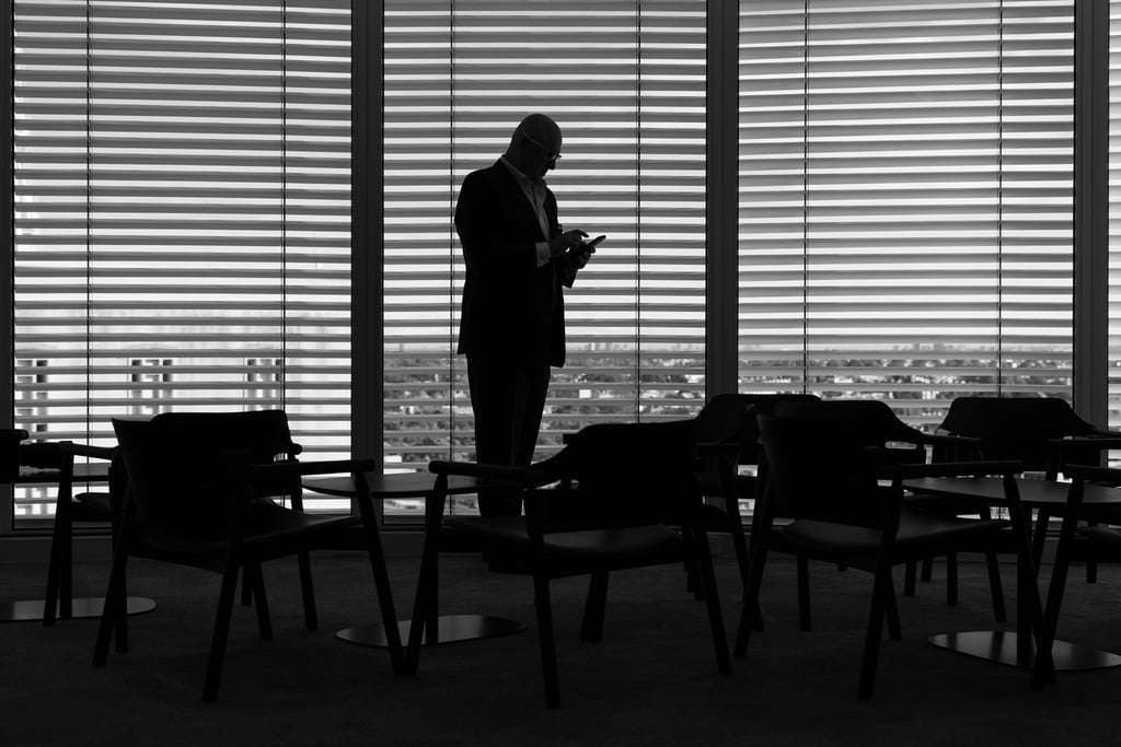 Silhouette of Nicholas using his mobile, standing in front of a window with blinds.