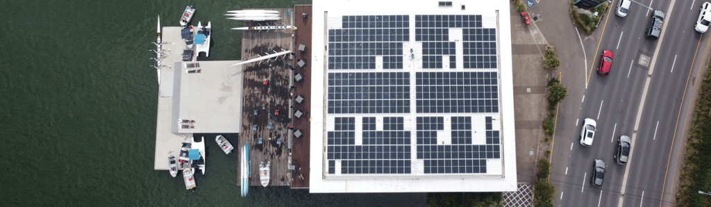 Aerial view of a building with a solar panel-covered roof next to a dock on a body of water. Several boats and cars are visible nearby on a road.