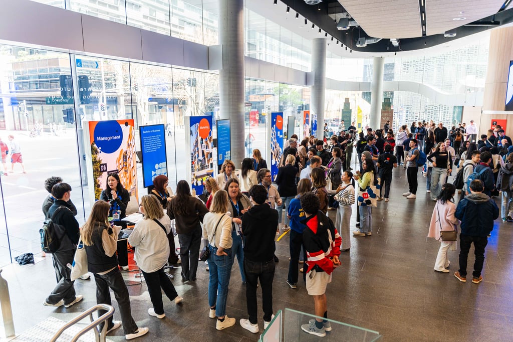 A crowd of students move about the floor of Building 2 as they speak with academics and students in front of large banners that read "Marketing", "Management" and "Finance".