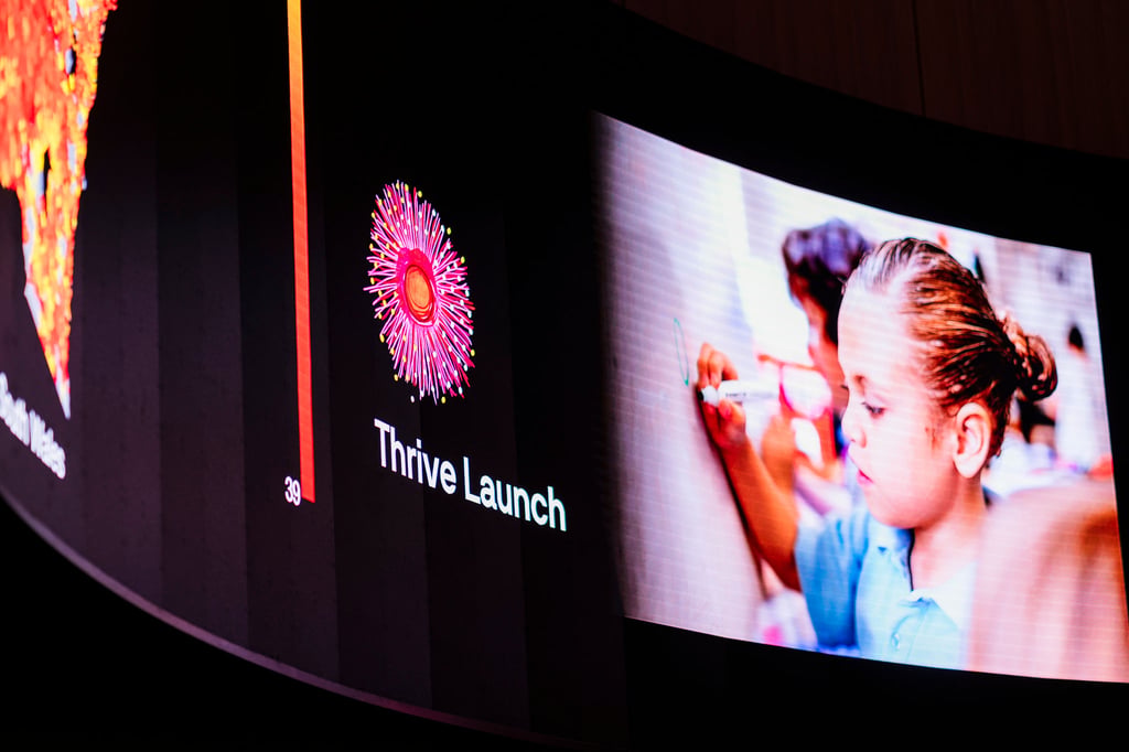 Large curved digital display at an event venue showing the words “Thrive Launch” beside a colourful circular graphic, with a photo of a child drawing on a wall displayed on the screen. The display is illuminated against a dark background.