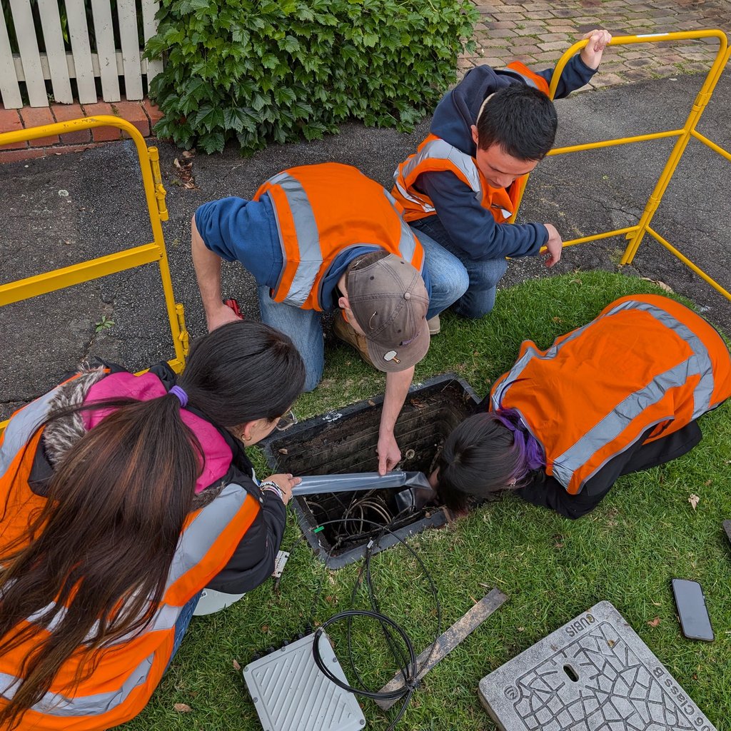 Four people wearing orange safety vests work on a maintenance hole on a grassy area. They are focused, suggesting teamwork and problem-solving. Yellow barriers surround them.