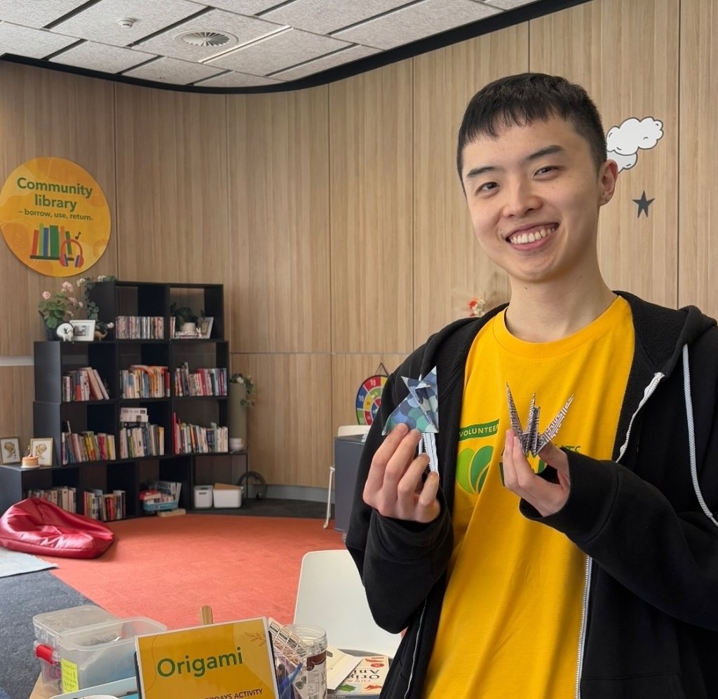 Volunteer holding origami creations beside an origami activity table in a campus community space with bookshelves and a community library in the background.