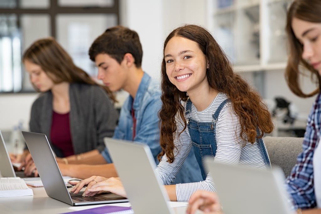Students seated at a table using laptop computers in a classroom or study space. One student in the centre looks toward the camera while other students focus on their work. Shelving and large windows are visible in the background.