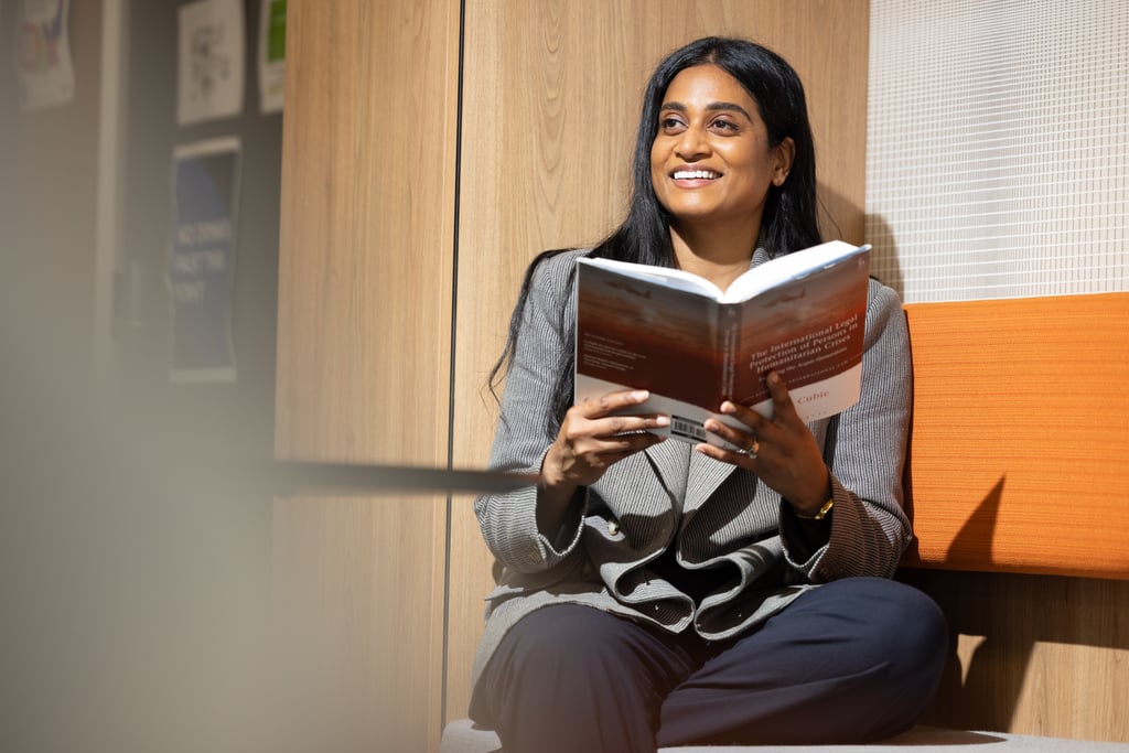 Ash in a grey blazer sitting on an orange couch reading a book. She looks up, smiling