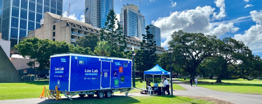 Mobile "Loo Lab" trailer in a park under a bright blue sky. It's next to a small blue canopy tent with people. Tall buildings and trees are in the background.