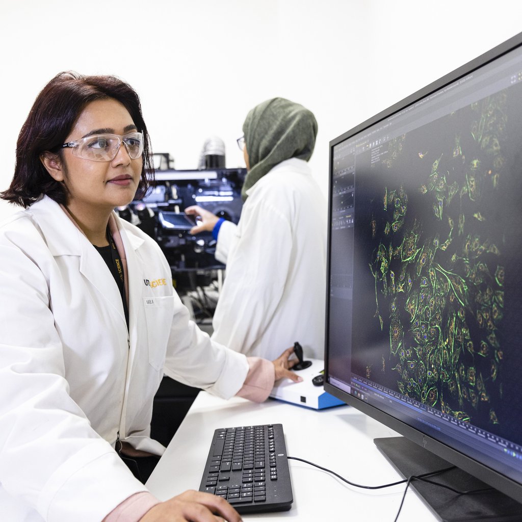 Two scientists in a lab, one analysing data on a computer screen showing green-tinted cellular images. Both wear lab coats and safety glasses, focused and professional.