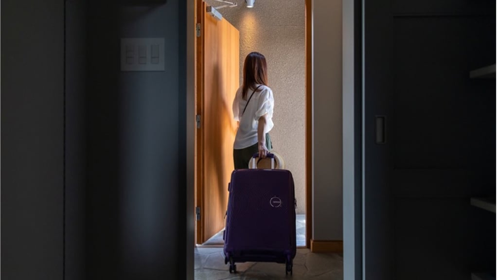 A woman seen from behind is leaving through an open door, pulling a purple suitcase.