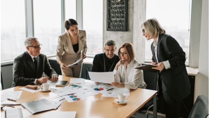 A group of five professionals in a modern office huddle around a table reviewing documents and charts. They appear focused and engaged in discussion.