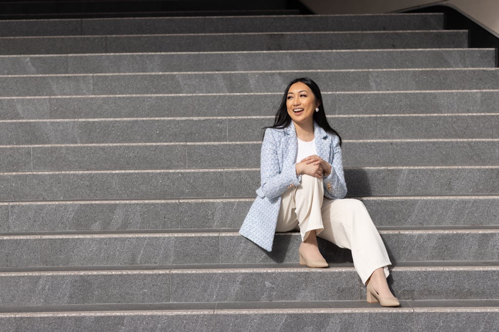 Arhita, wearing a light blue blazer and beige trousers, sits on steps, laughing