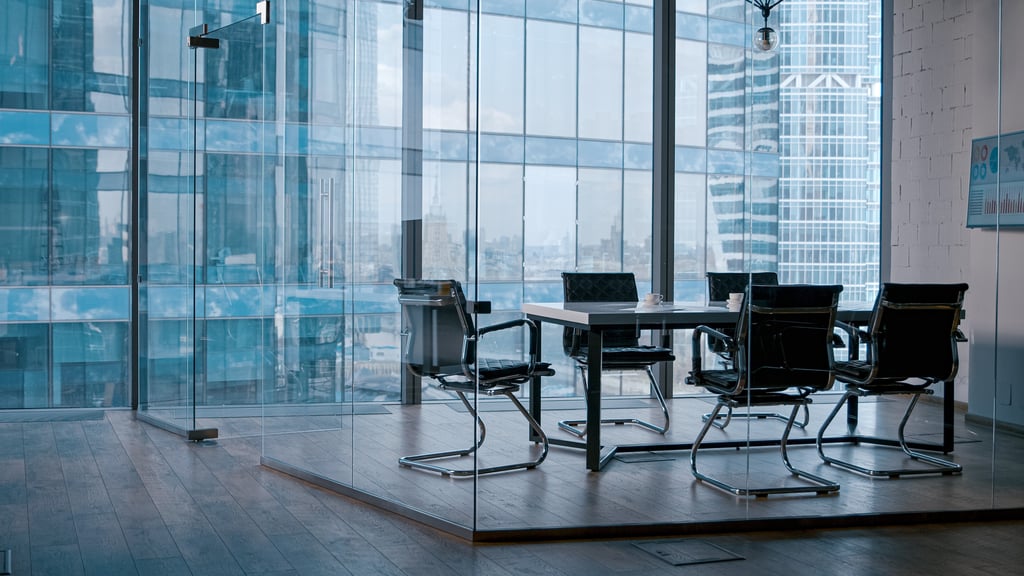 Empty meeting room in office, six black leather chairs are placed around a glass table while skyscrapers can be viewed in the background.