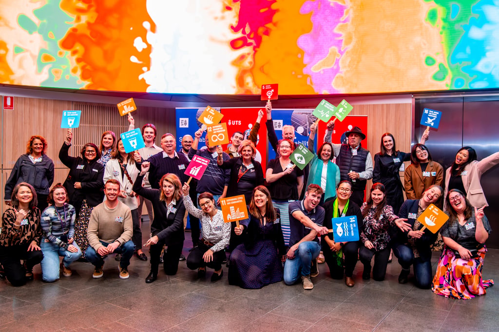 Group of people posing indoors at UTS Global Goals Month, while holding colourful signs representing the United Nations Sustainable Development Goals.
