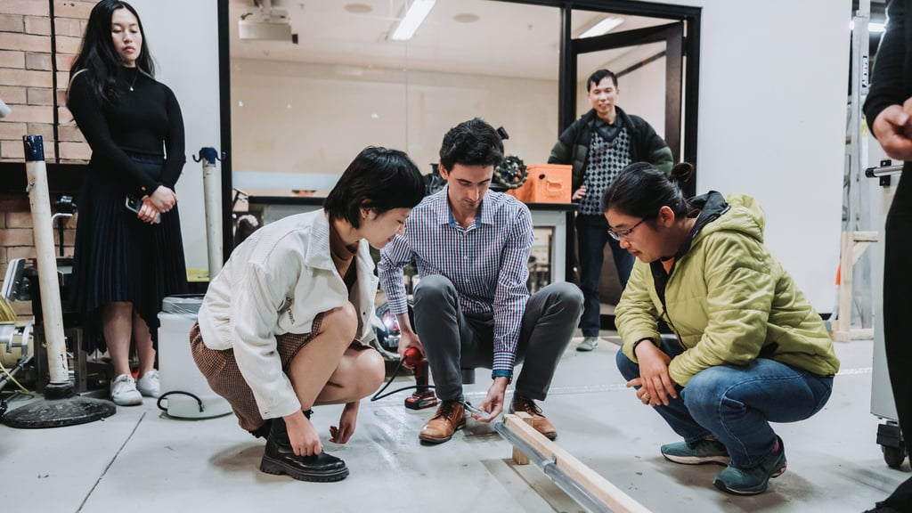 A group of people in a workshop focus intently on a woodworking project. Three crouch on the floor, while two stand nearby, observing attentively.