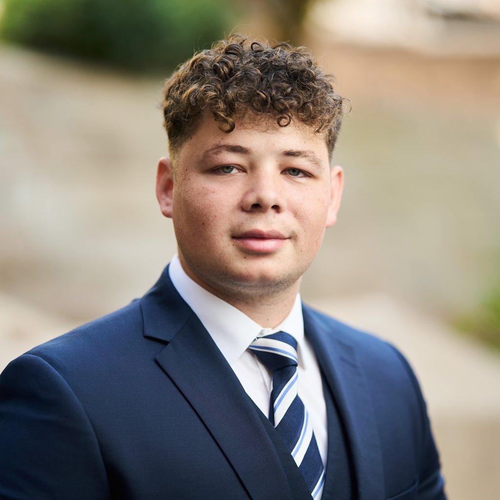 Deon wears a dark blue suit, white shirt and striped blue and white tie