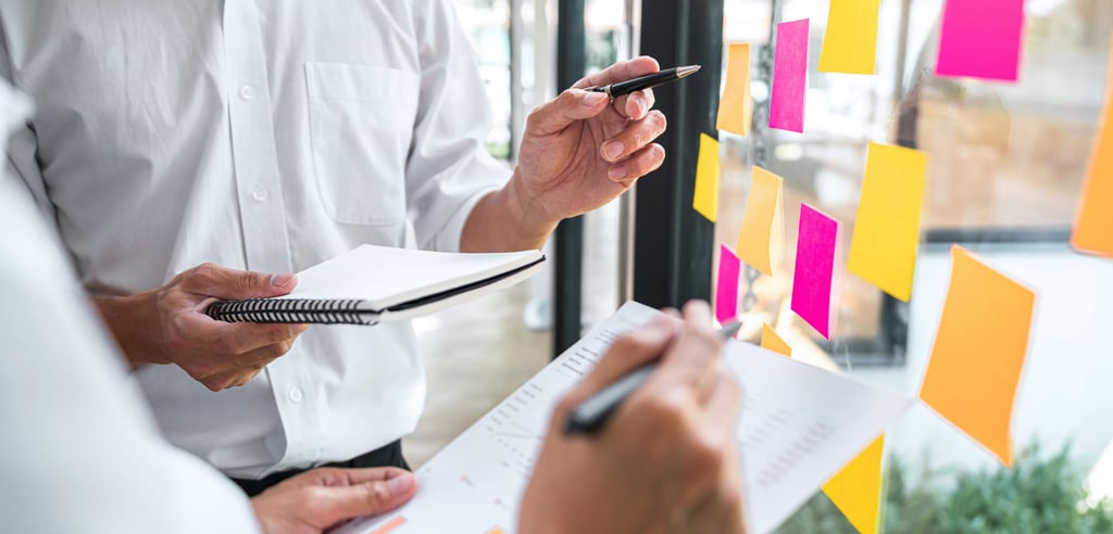 Two people collaborate in an office, holding notebooks and pens. Bright sticky notes cover a glass wall, conveying a focused, brainstorming atmosphere.