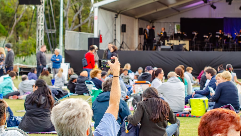 A raised hand holding a phone captures the crowd gathered at an outdoor live music event in a public park, people sitting in front of a performance stage. A lively community atmosphere in Australia. Picture: Doublelee/Adobe Stock