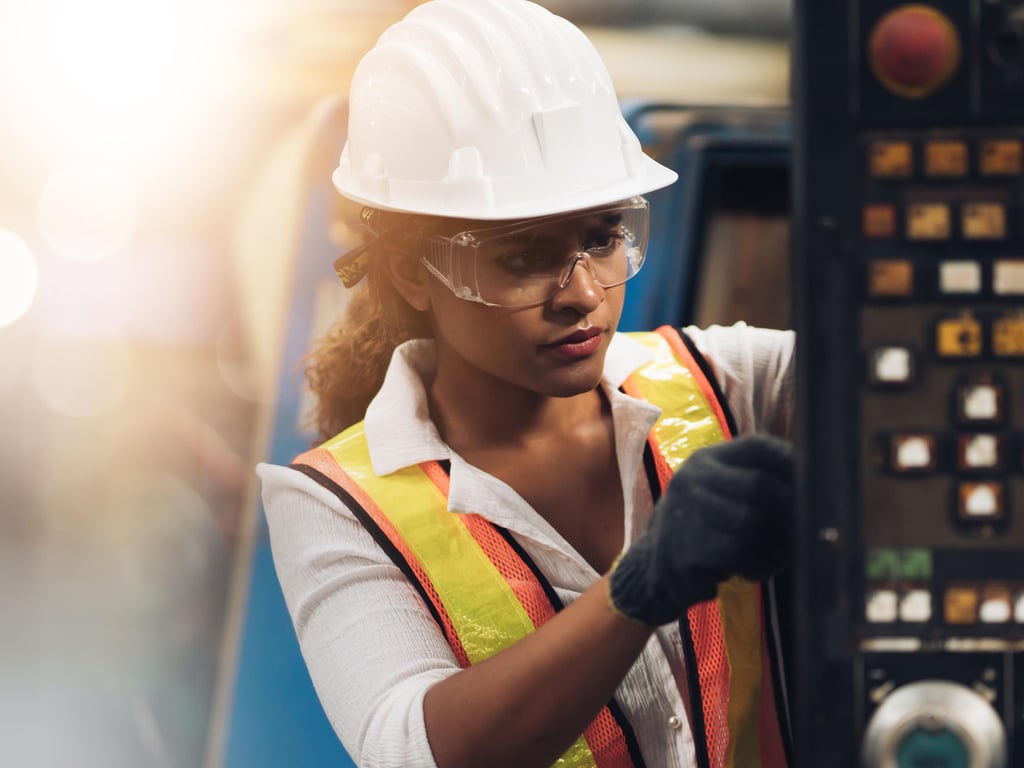 A focused female engineer in a hard hat and safety vest operates machinery, examining controls with precision. Bright industrial background.