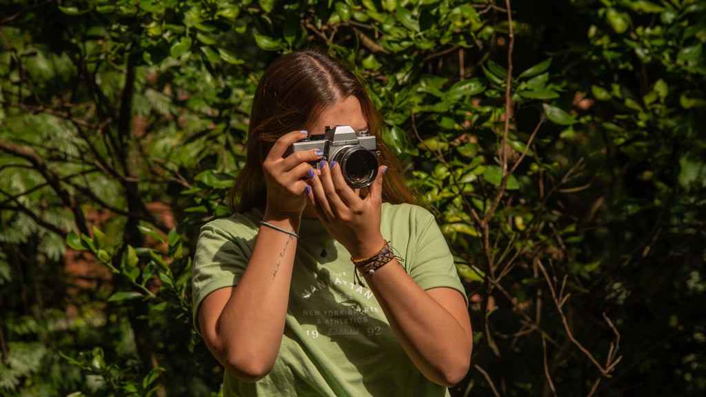 A person in a green shirt holds a vintage camera, surrounded by lush greenery. Sunlight filters through the leaves, creating a serene, focused atmosphere.