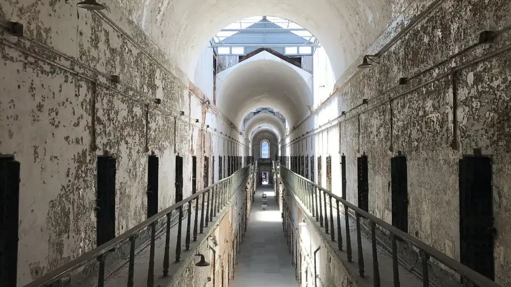 Interior of a historic prison cell block with peeling white walls, rows of dark cell doors on both levels, and a long central walkway leading to an arched window at the far end.