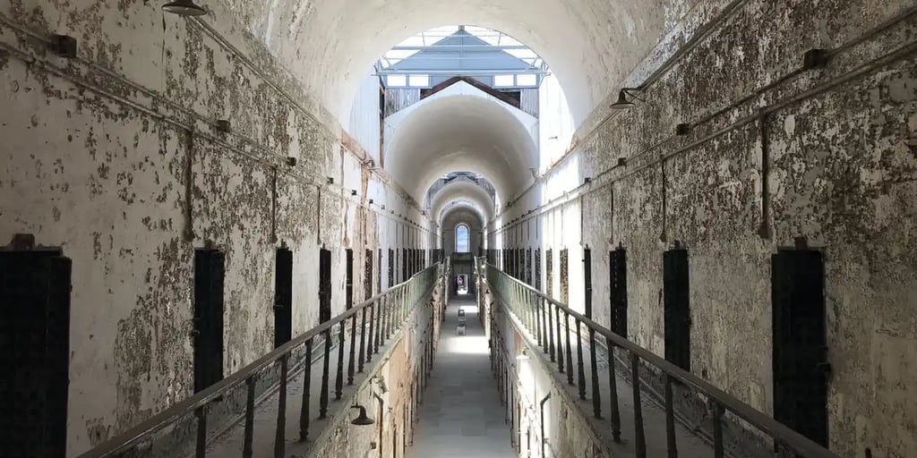 Interior of a historic prison cell block with peeling white walls, rows of dark cell doors on both levels, and a long central walkway leading to an arched window at the far end.