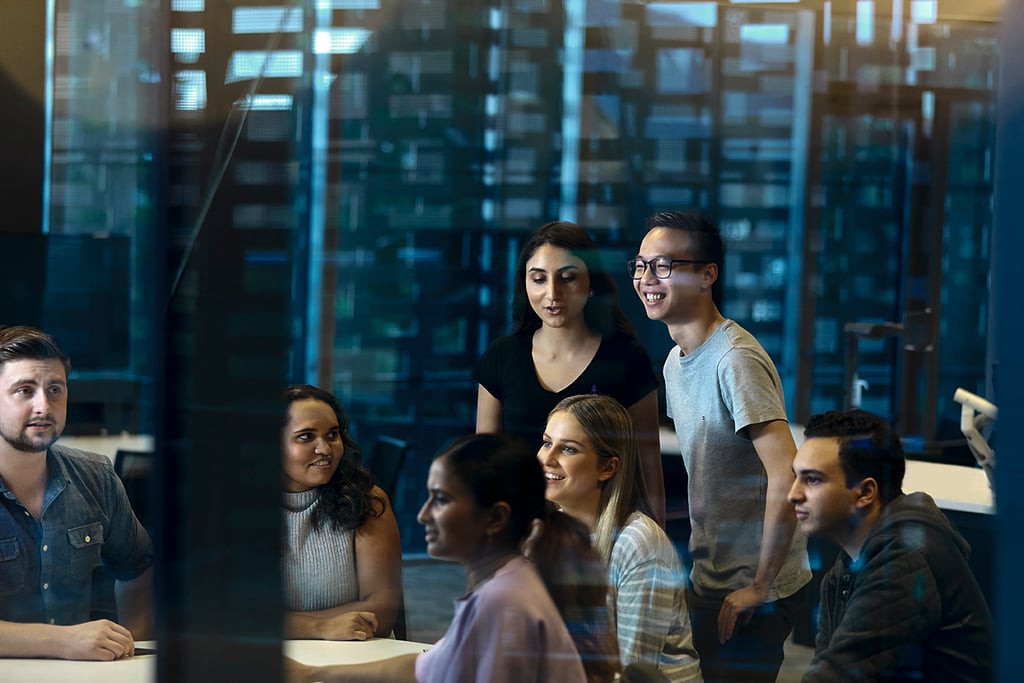 A group of people collaborating around a table in a modern office, viewed through a glass wall.