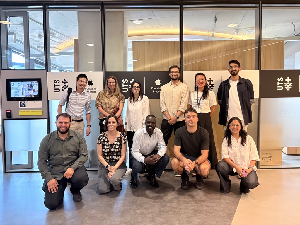 A diverse group of eleven people, smiling, poses in front of a glass wall with "UTS" logos. Some are standing, others kneeling, in a modern office setting.