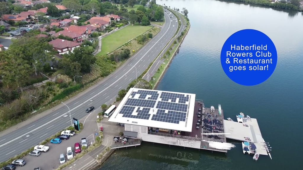 Aerial view of the Haberfield Rowers Club and Restaurant located beside a river, with a large solar panel installation covering the rooftop. A road runs alongside the waterfront venue, with residential homes and trees on the opposite side. The image includes the text “Haberfield Rowers Club & Restaurant goes solar!” within a blue circular graphic.