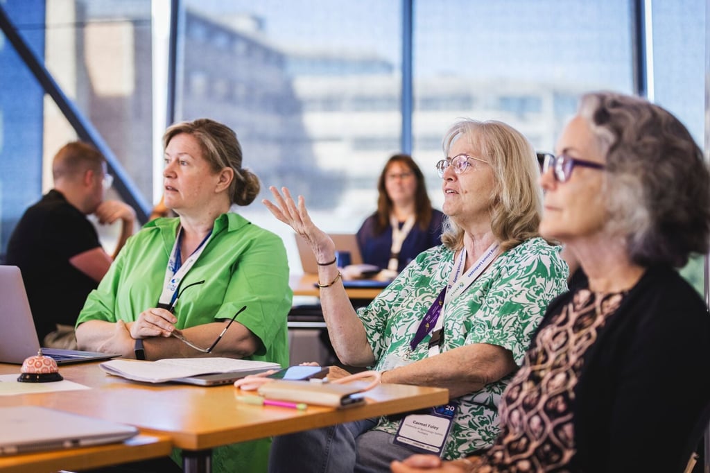 Participants seated around a table during a workshop, listening to a speaker and contributing to discussion, with laptops, notebooks and conference lanyards visible.