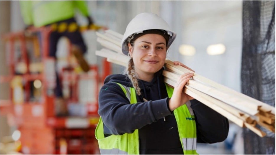 A woman in a construction site smiles while carrying wooden planks. She wears a white hard hat and a reflective vest, exuding confidence and teamwork.